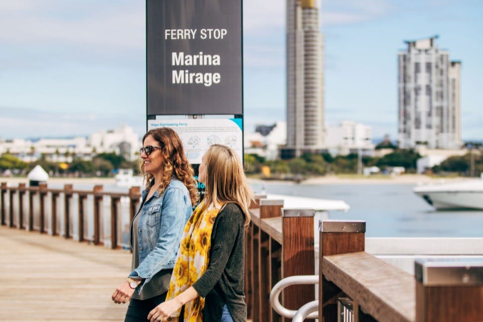 Две женщины идут по набережной возле знака «Паромная остановка Марина Мираж» с городским пейзажем на заднем плане.
