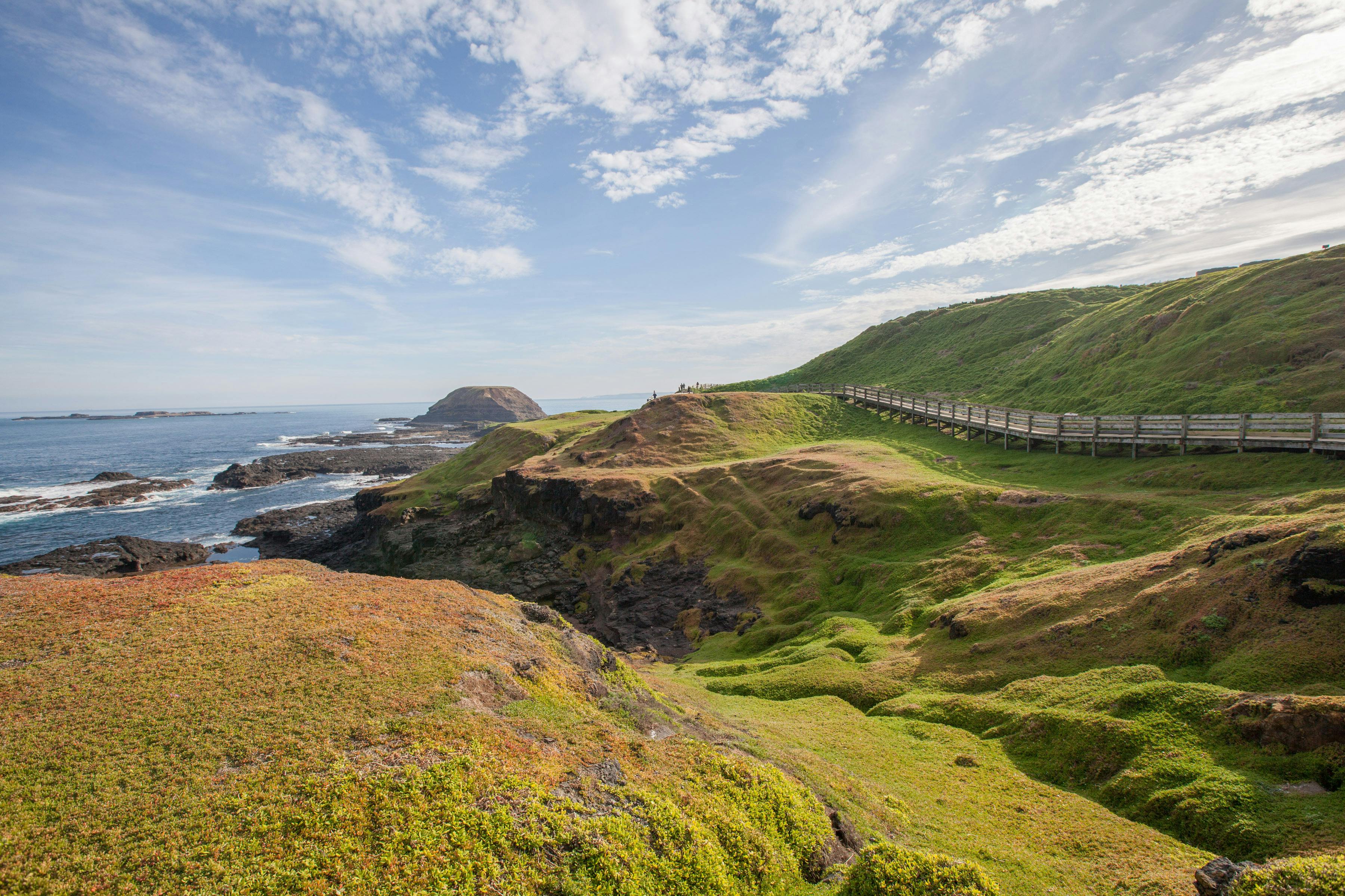 Scenic coastal landscape with green rolling hills, cliffs, and a wooden path under a partly cloudy sky, extending towards the ocean.