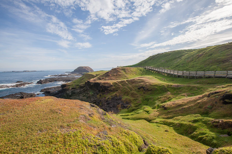 Scenic coastal landscape with green rolling hills, cliffs, and a wooden path under a partly cloudy sky, extending towards the ocean.