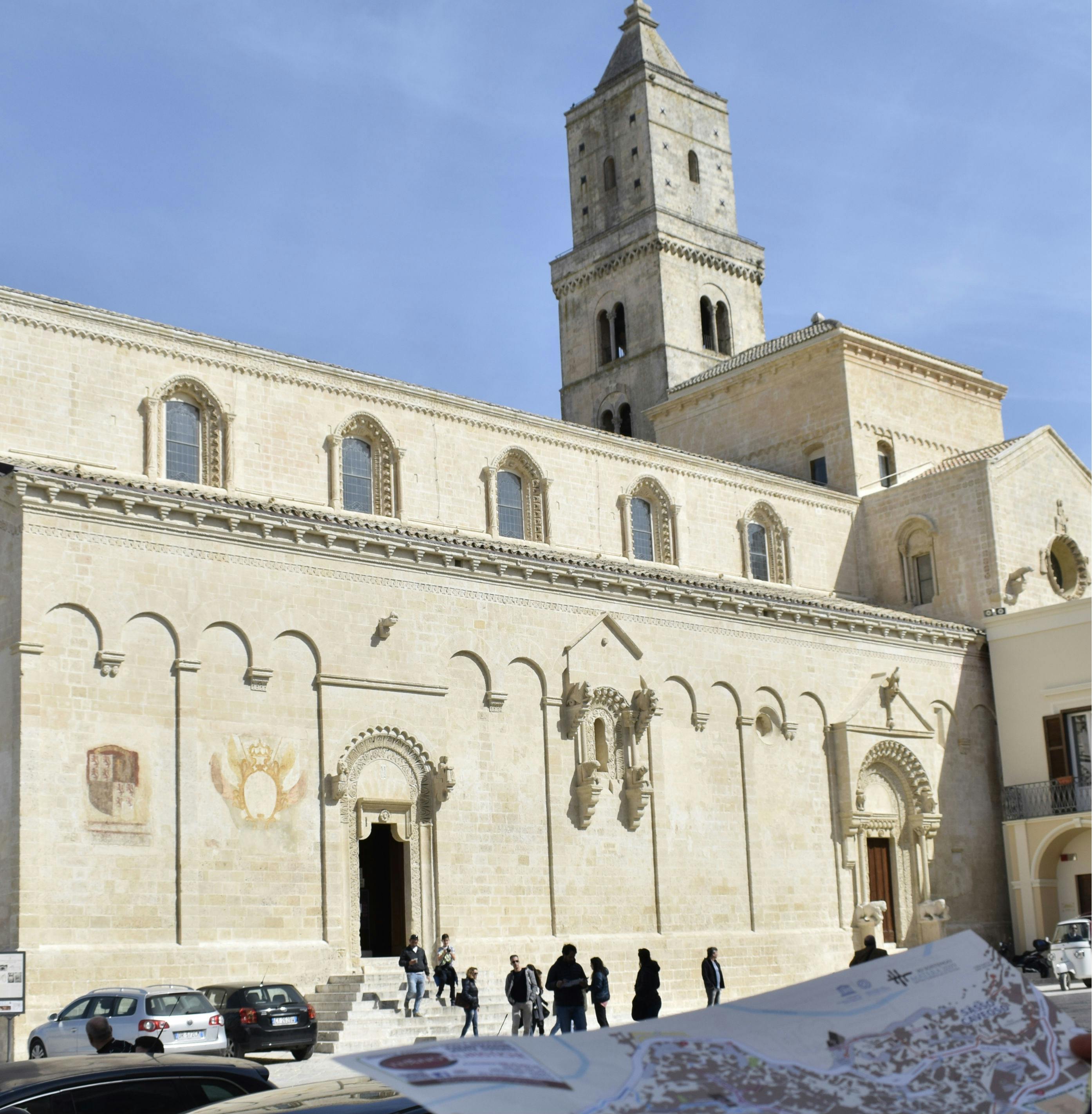 Een groep mensen staat voor een historische kalkstenen kerk met boogramen en een hoge toren onder een strakblauwe hemel.