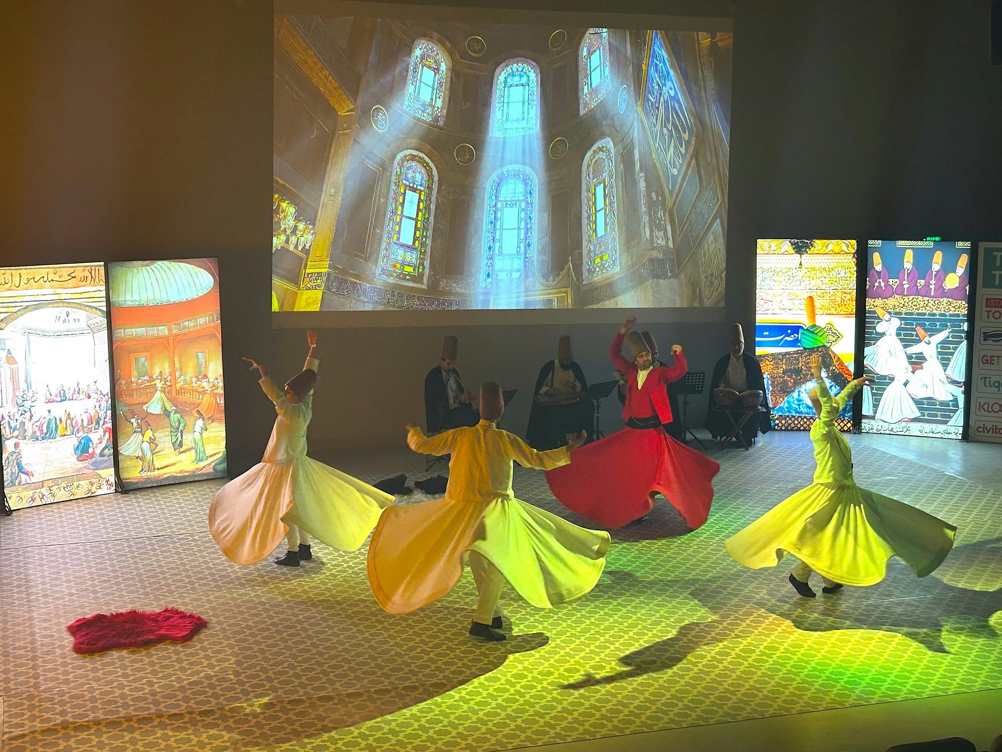 Χορευτές με πολύχρωμα περιστρεφόμενα κοστούμια εκτελούν με φόντο προβαλλόμενες εικόνες σε ένα αμυδρά φωτισμένο δωμάτιο.