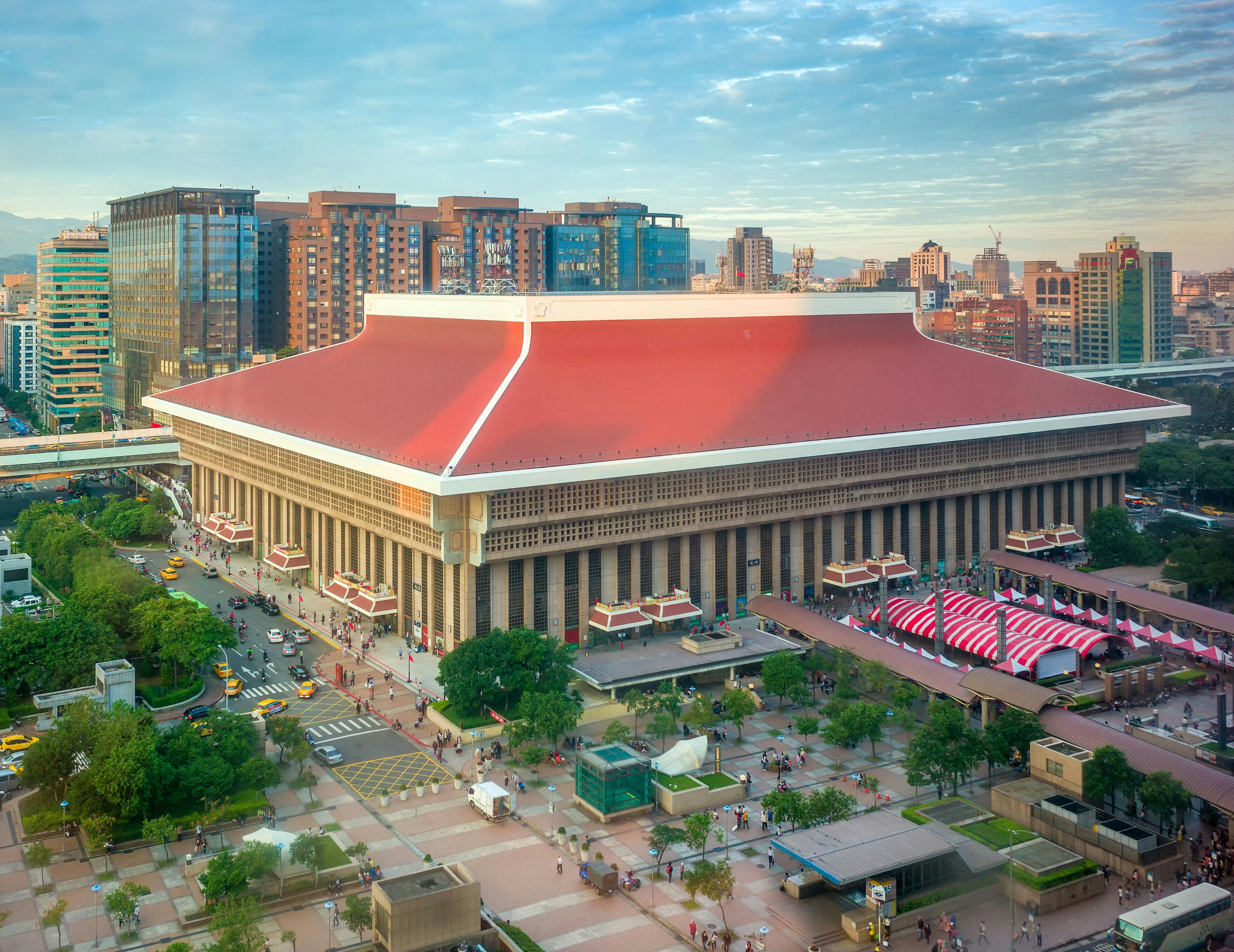 Velká budova s červenou střechou a složitou fasádou je obklopena stromy, lidmi a taxíky v městské oblasti.