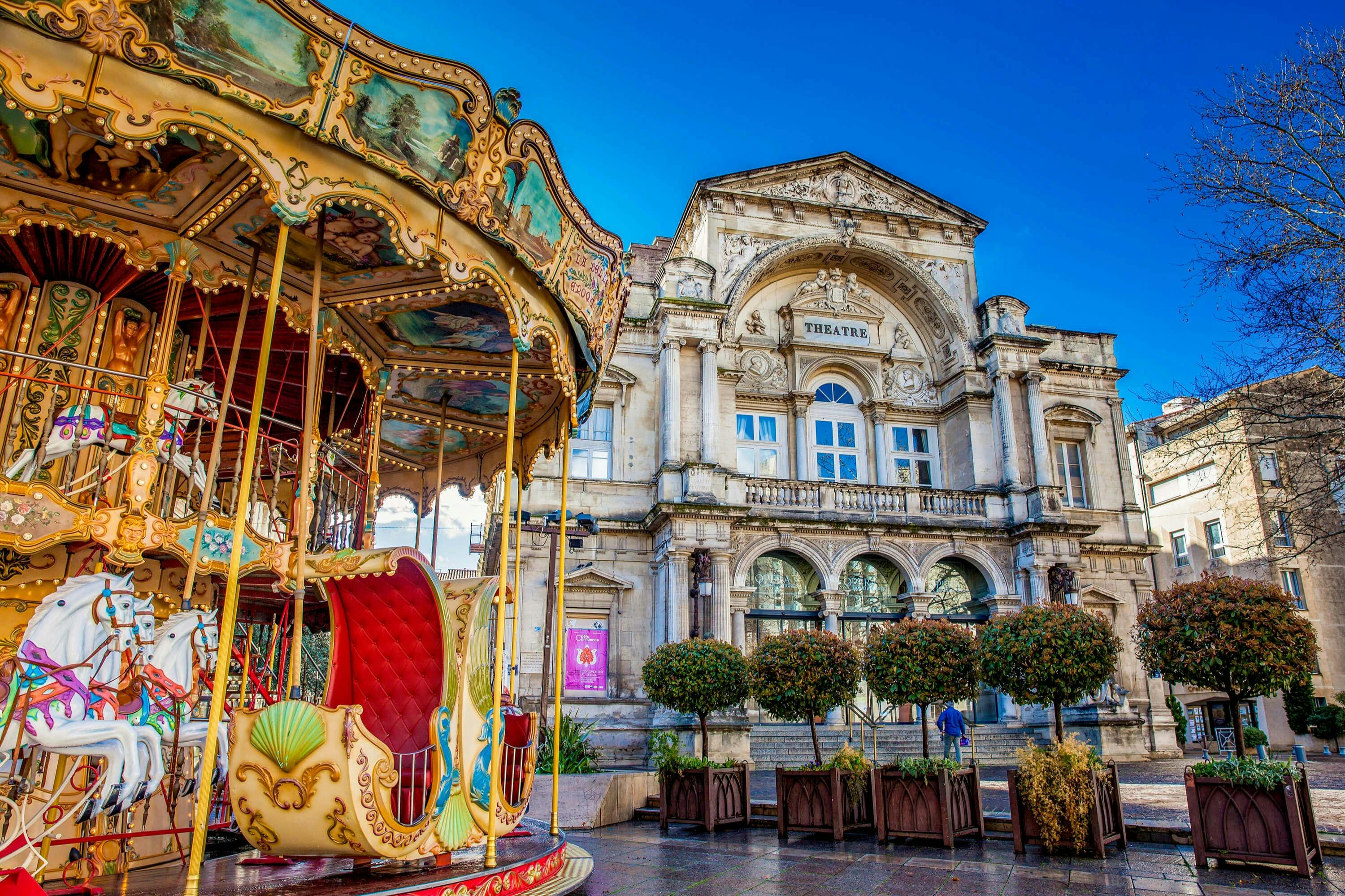 A colorful carousel with ornate details stands in front of a historic theater building under a clear blue sky.