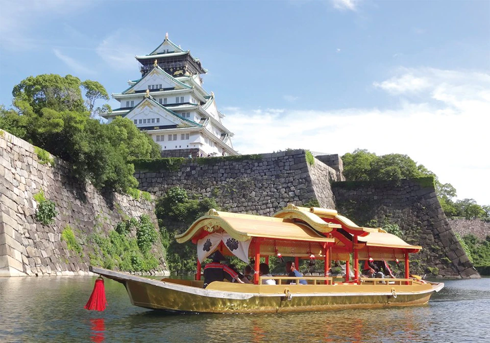 A traditional Japanese boat with a golden roof floats on water, with a historic castle and stone walls in the background.