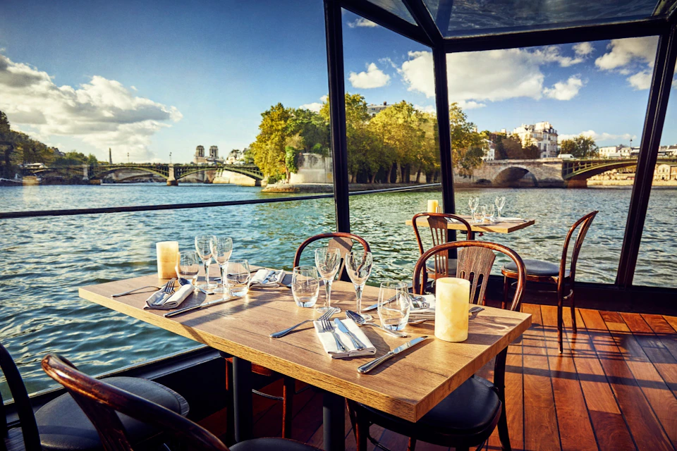 Dinner tables set with glassware and candles on a boat with large windows, overlooking a river and a bridge.
