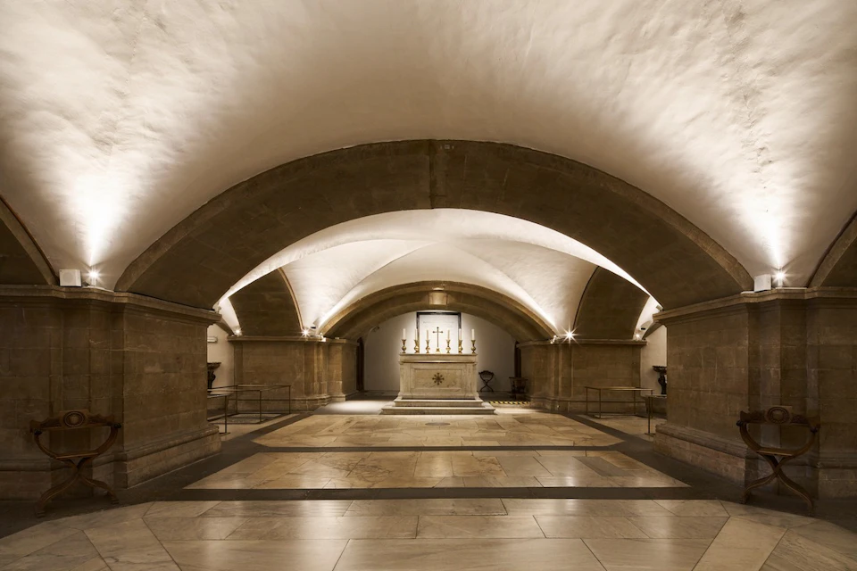 A vaulted stone crypt with an altar and cross at its center, illuminated by warm lighting.