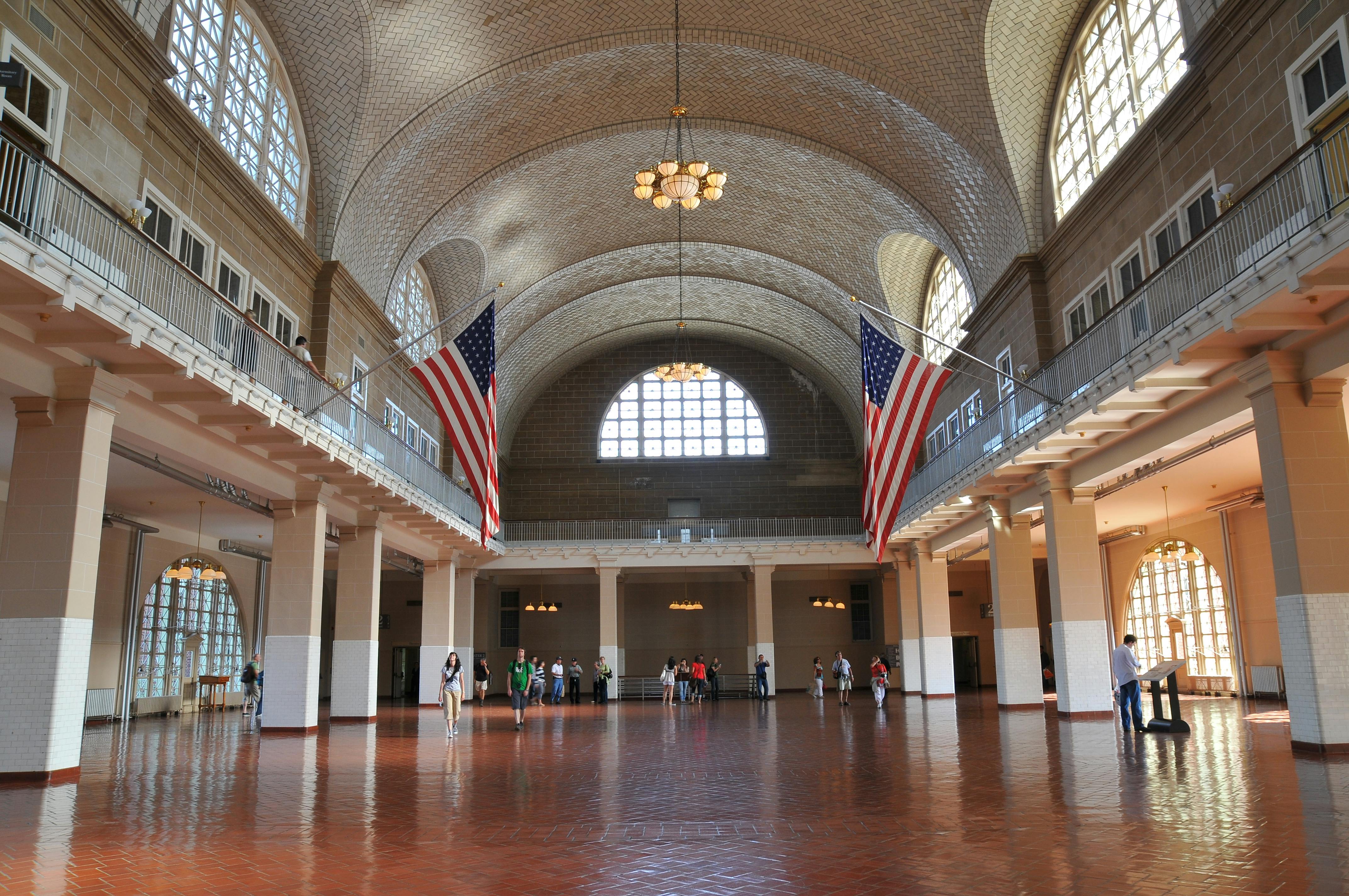 아치형 천장, 커다란 창문, 두 개의 미국 국기, 샹들리에, 타일 바닥을 걷는 사람들이 있는 그랜드 홀입니다.