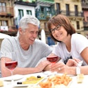 Una coppia di anziani sorridenti con occhiali che si godono un pasto e un drink all'aperto, con edifici colorati sullo sfondo.