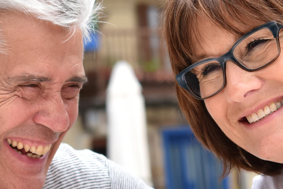 A smiling older couple with glasses enjoying a meal and drinks outdoors, with colourful buildings in the background.