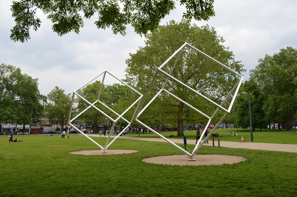 Two large metal cube sculptures in a park with lush green grass, trees, and people sitting or walking in the background.