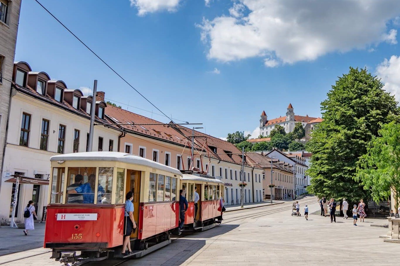 Κόκκινα τραμ σε έναν δρόμο με ανθρώπους σε μια ευρωπαϊκή πόλη. Στο βάθος βρίσκονται κτίρια με κόκκινες στέγες και ένα κάστρο σε λόφο.