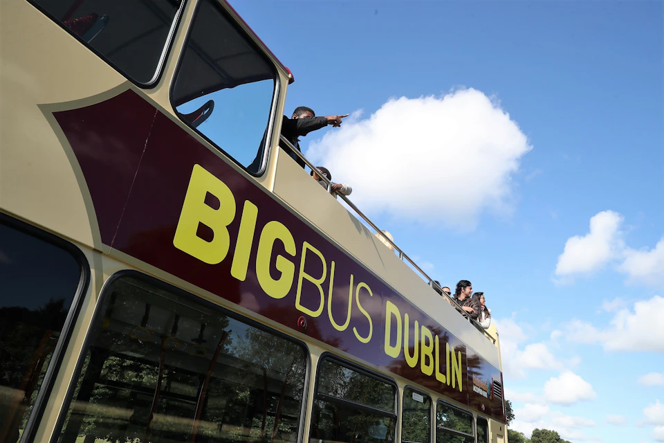 Tourists ride on the top deck of a "Big Bus Dublin" double-decker under a blue sky with a white cloud.