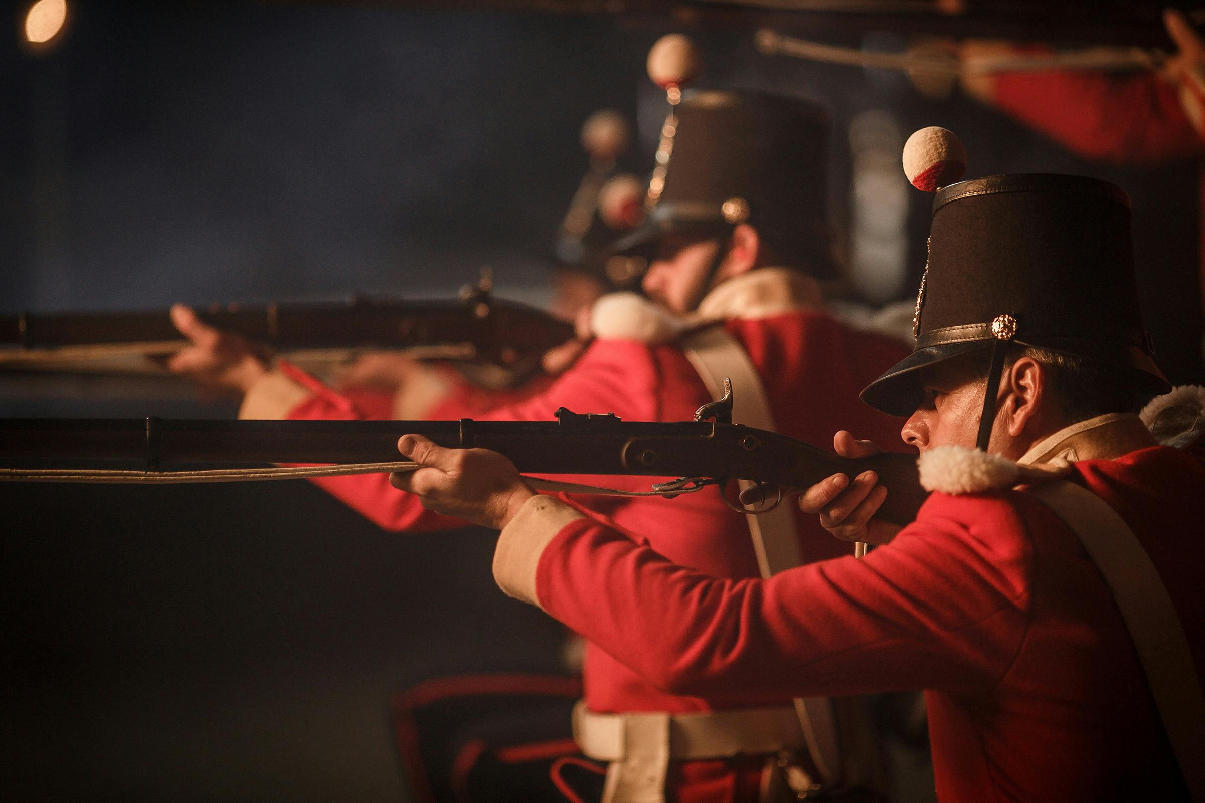 Soldiers in red and white uniforms aim rifles, wearing black hats with white and red pom-poms in a dark, smoky setting.