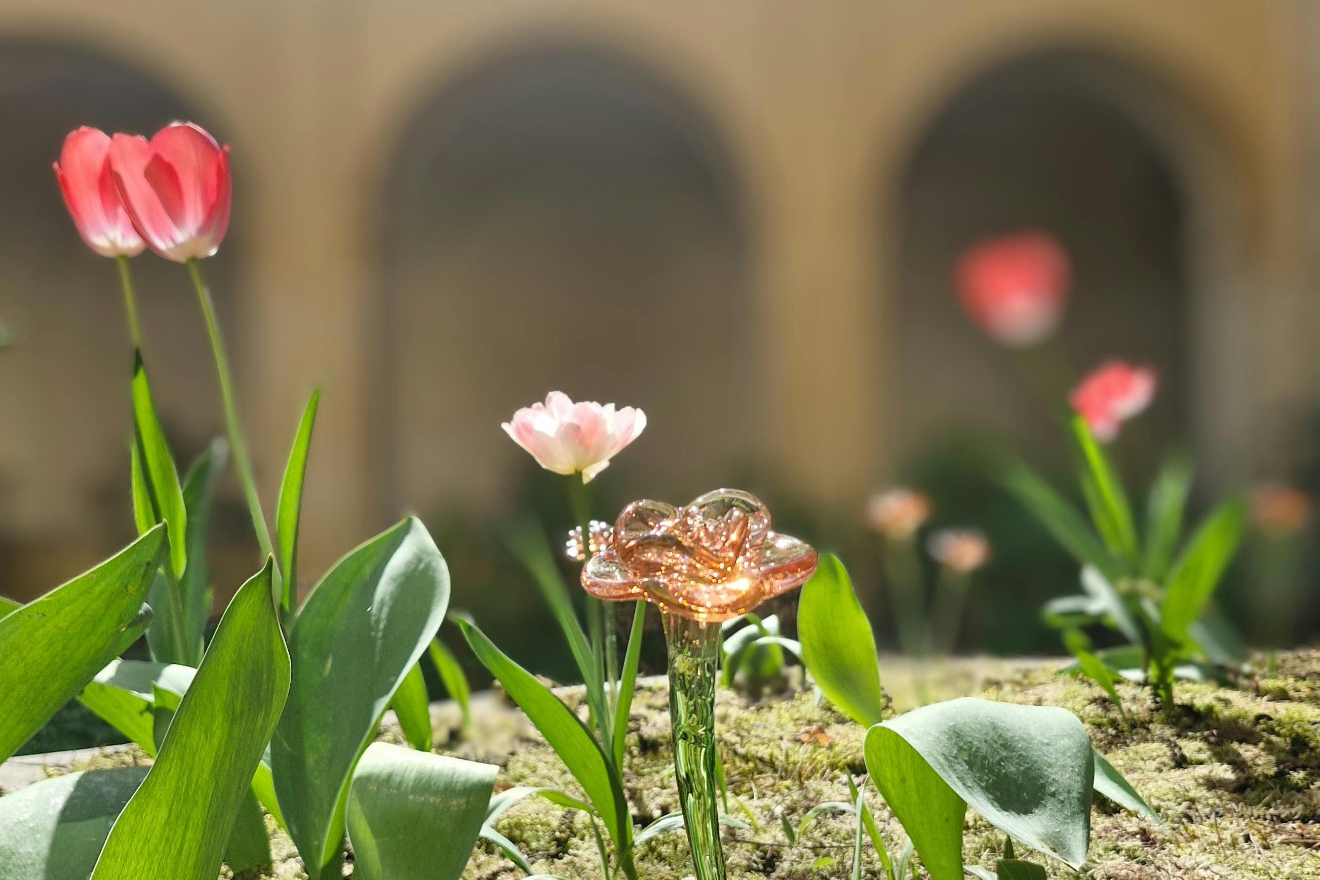 A metallic rose sculpture amidst green leaves and blooming pink flowers with an arch in the blurred background.