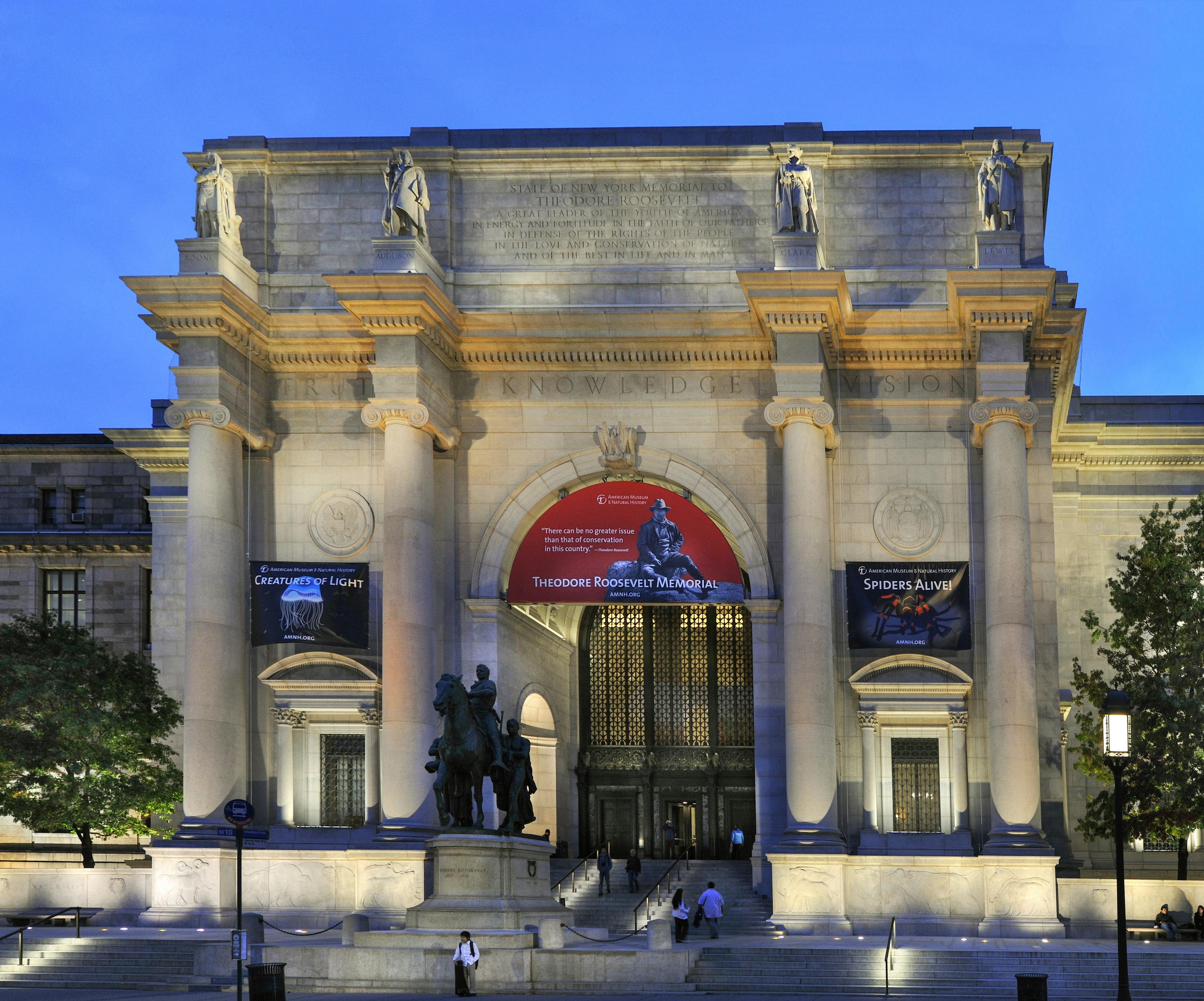 Large, neoclassical building entrance with columns, banners, statues, and steps. People walk at the base under a twilight sky.