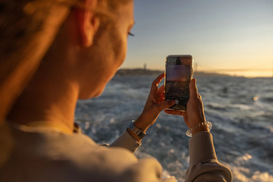 A person takes a photo of the ocean and distant shoreline with a smartphone at sunset.