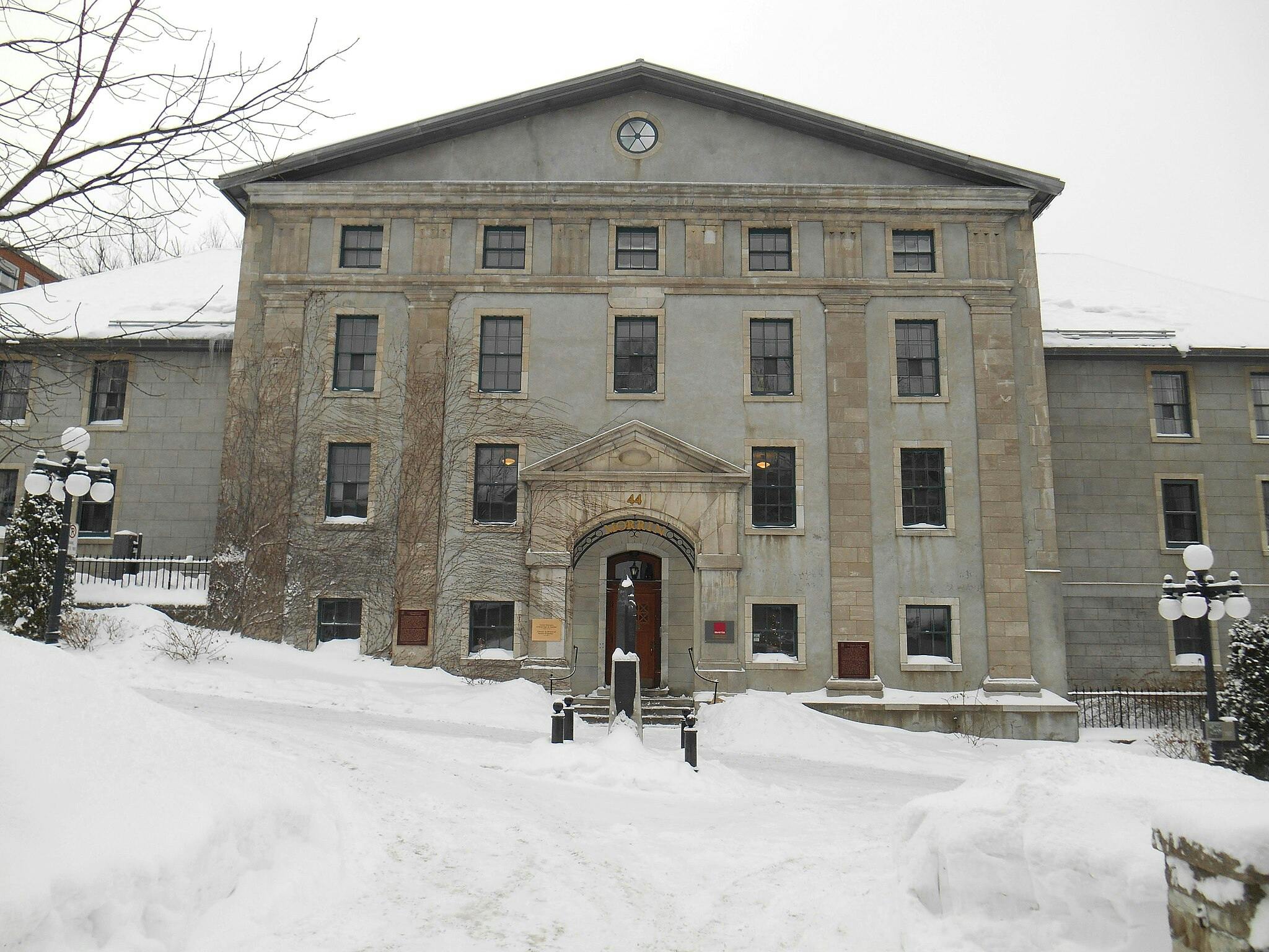 Velká šedá kamenná budova pokrytá sněhem, s holými stromy a pouličními lampami vpředu a cestičkou vedoucí ke vchodu.