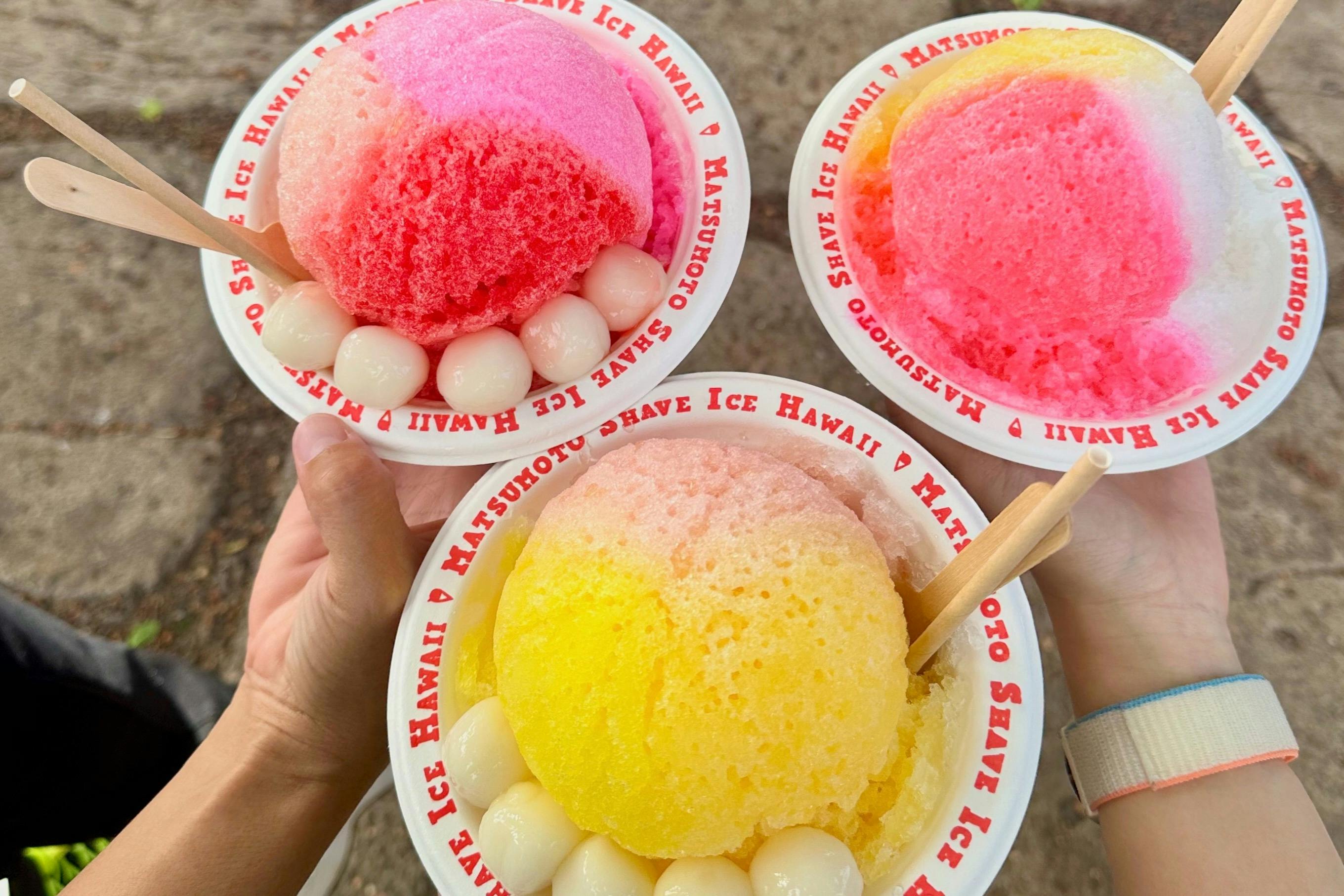 Three colorful shave ice bowls with toppings, held by hands, labeled "Matsumoto Shave Ice Hawaii" on the rim.