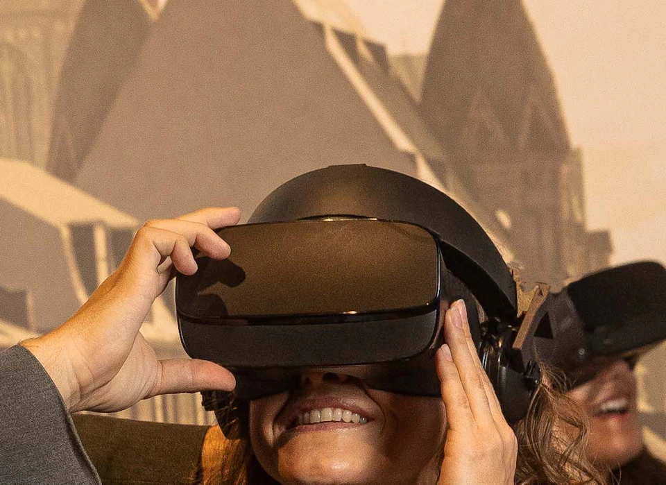 A man in historical attire helps a seated woman wearing a VR headset inside a themed setting with a mural of buildings.