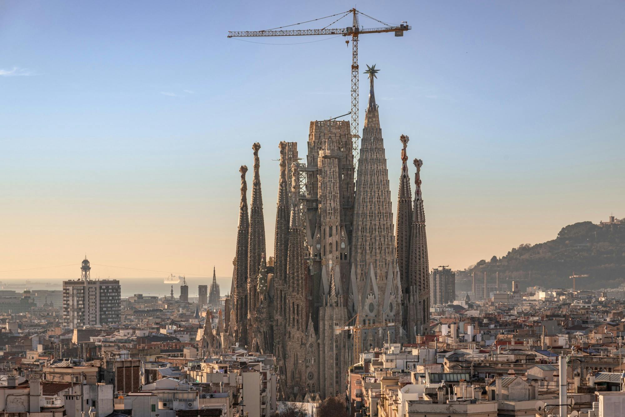 Die im Bau befindliche Sagrada Familia mit Kränen vor einer Stadtkulisse bei Sonnenuntergang in Barcelona.