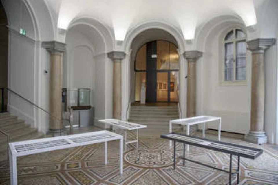 Interior view of an exhibition space with arched ceilings, columns, and tables displaying documents. Stairs and artwork in the background.
