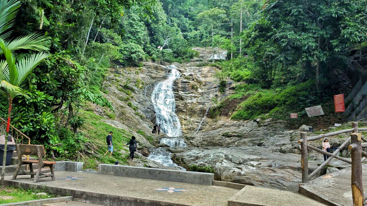 A waterfall cascades down rocky terrain surrounded by lush green trees; people are walking and exploring near its base.