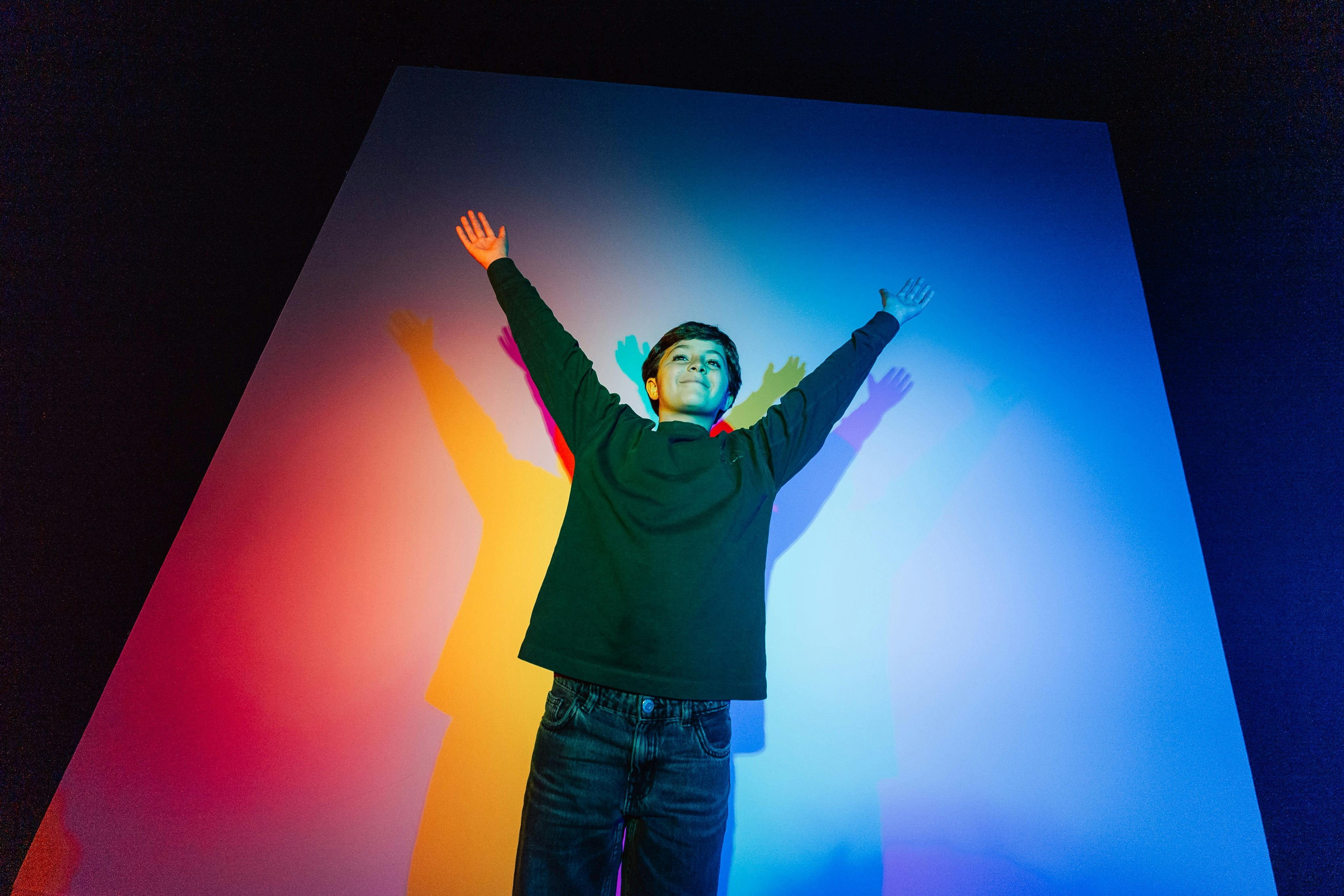 A person stands with arms outstretched against a wall illuminated by colorful lights casting vibrant shadows.