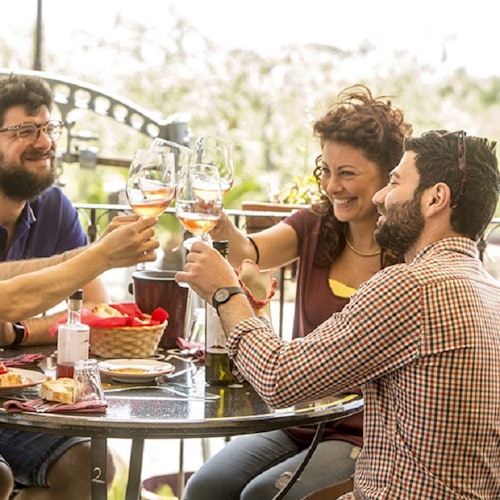 Three people toast with wine glasses around a table with food at an outdoor dining area, all smiling and enjoying each other’s company.