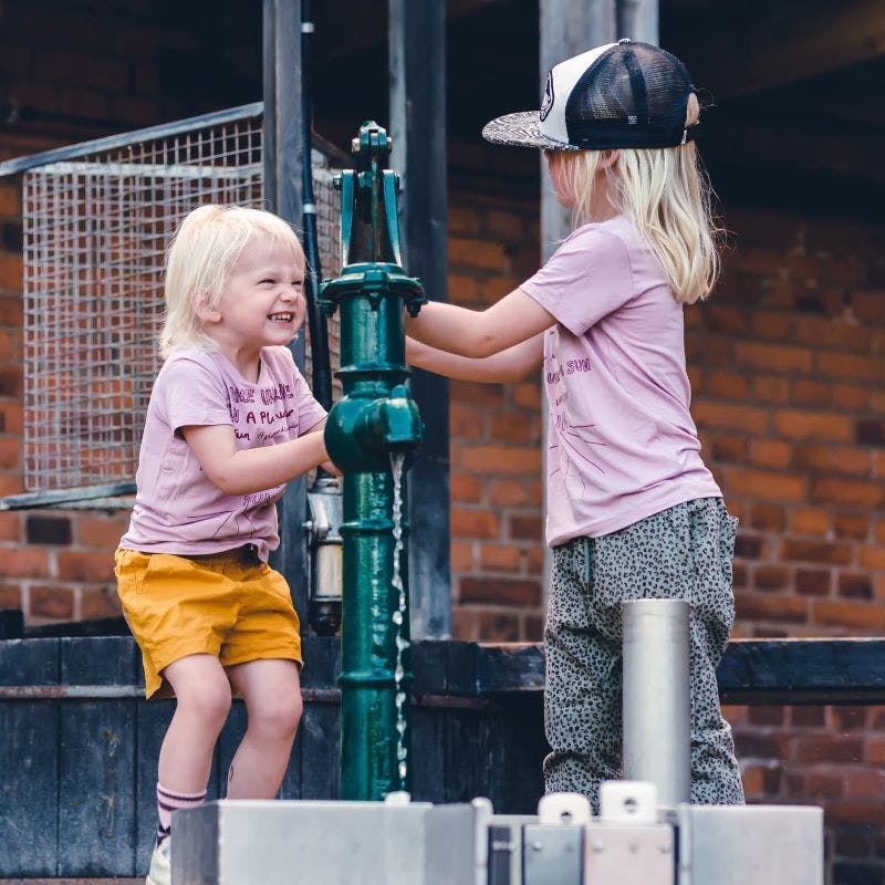 Två små barn i rosa skjortor leker med en grön vattenpump; en bär en keps, den andra ler ivrigt.