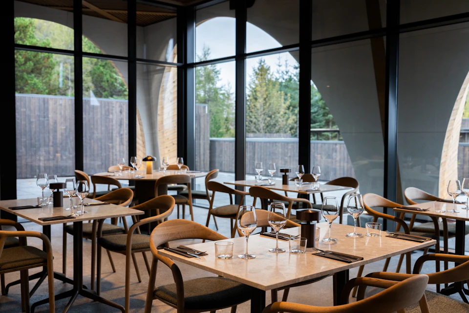 A modern restaurant interior with wooden chairs and tables set with glassware, overlooking an outdoor area through large windows.