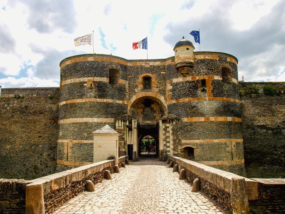 石造りの要塞入り口には2つの円形の塔があり、上には3つの旗が掲げられ、アーチ型の門に通じる石畳の通路があります。
