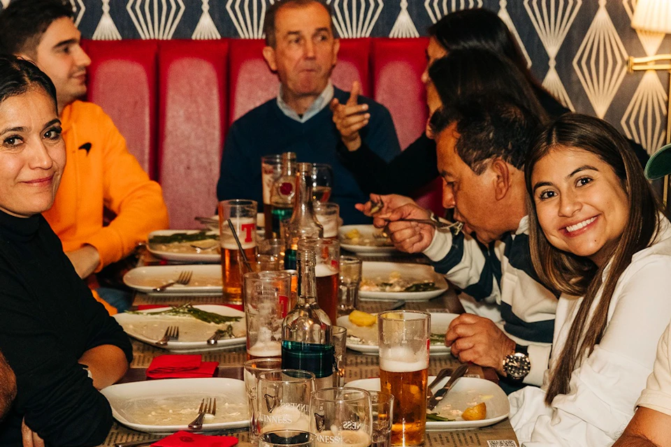 A group of people seated around a long dining table with plates of food, drinks, and a lit lamp in the background.