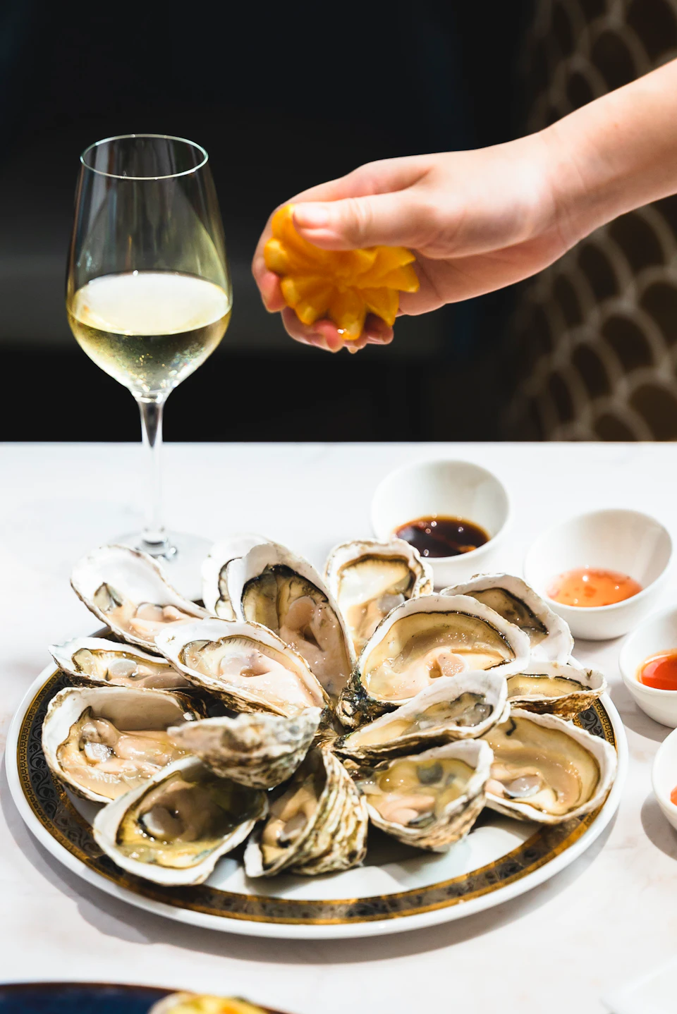 A plate of opened oysters with condiments and a glass of white wine, as a hand squeezes a lemon over the oysters.