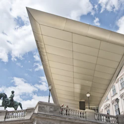 En ryttarstaty i brons och två personer på en balkong under ett modernt vinklat tak, mot en delvis molnig himmel.