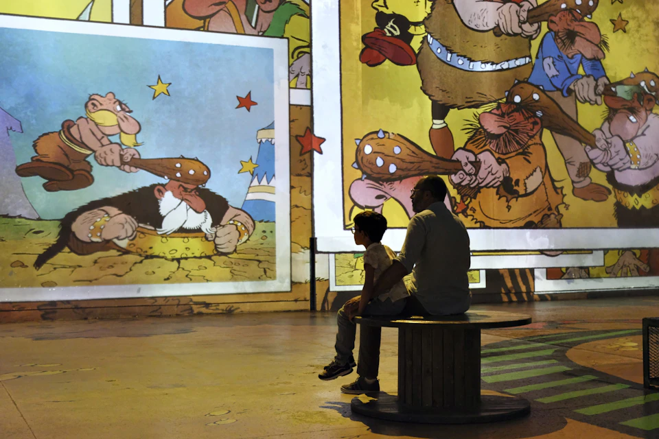 A child and an adult sit on a bench, looking at large colorful cartoon illustrations projected on a wall.
