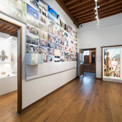 Museigalleri med fotovägg, glasmonter med artefakter och trägolv under takbjälkar.