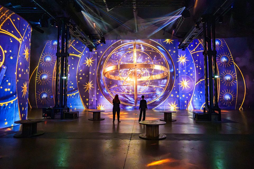 Two people standing in front of a large, illuminated globe design with star patterns. The scene is indoors with dramatic lighting.
