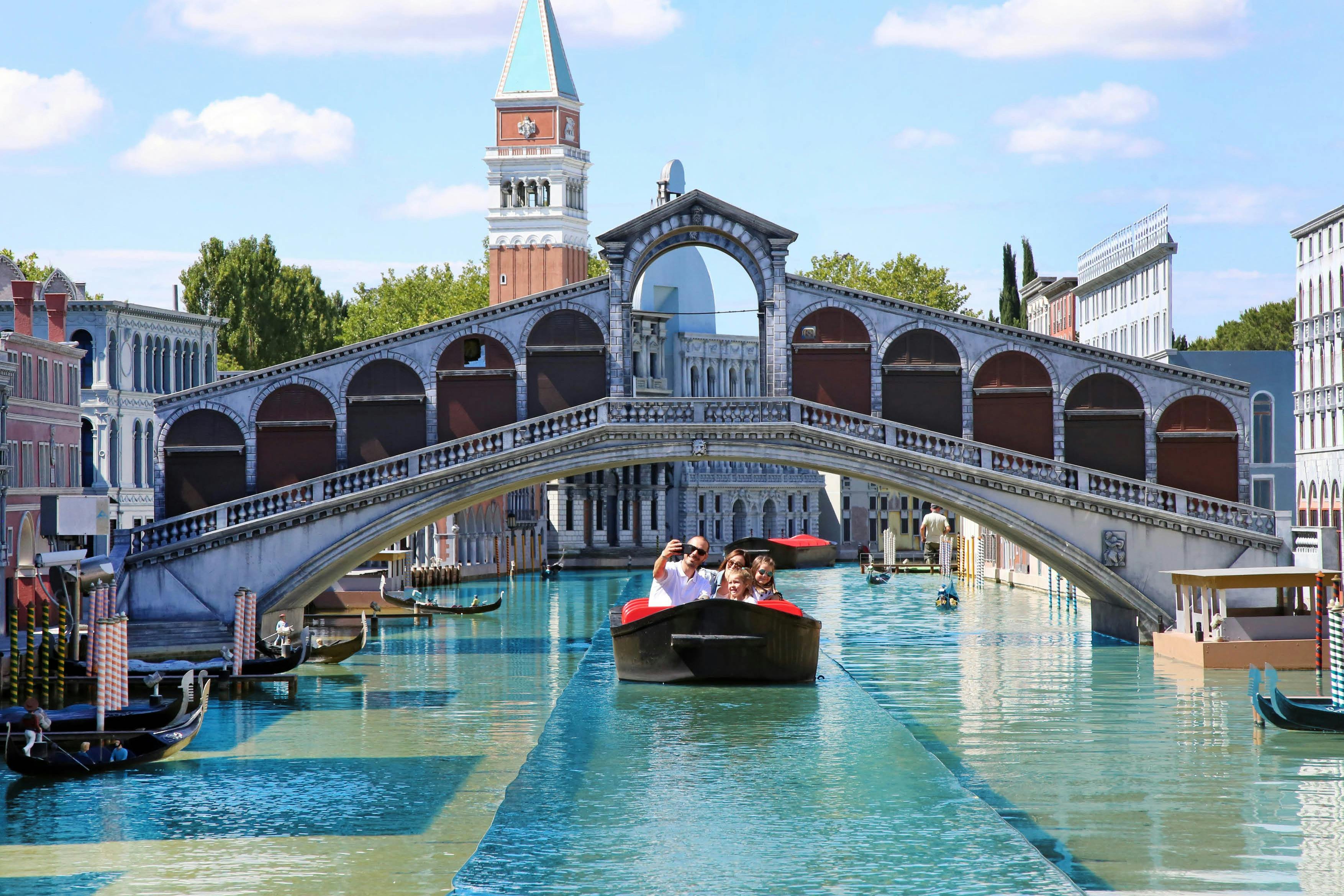 Eine Gruppe von Menschen genießt eine Gondelfahrt auf einem Kanal unter einer Brücke, mit Architektur, die an Venedig erinnert, im Hintergrund.