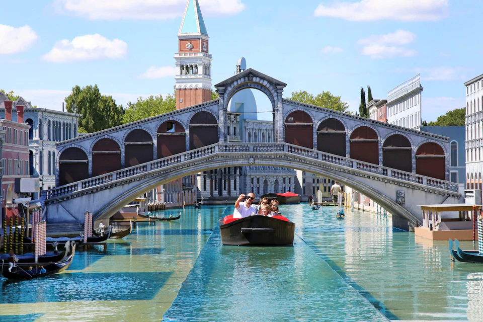 A group of people enjoy a gondola ride on a canal under a bridge, with architecture resembling Venice in the background.