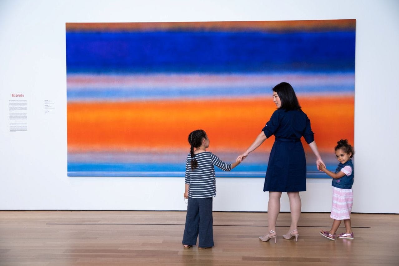 Una mujer cogida de la mano con dos niños delante de un gran cuadro abstracto con vibrantes tonos azules y naranjas.