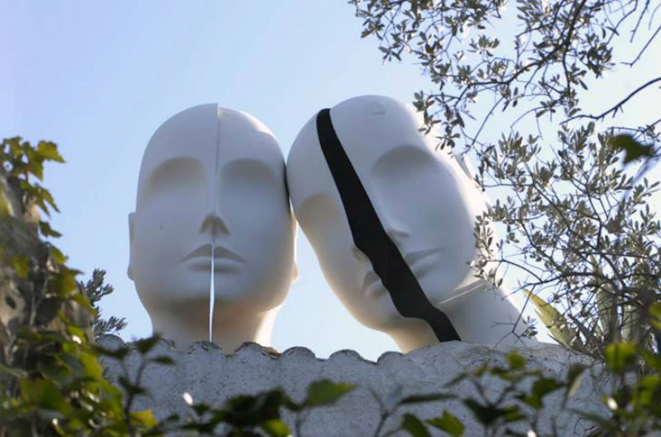 Two mannequin heads with split faces, one with a white stripe and the other with a black stripe, set against a blue sky and foliage.