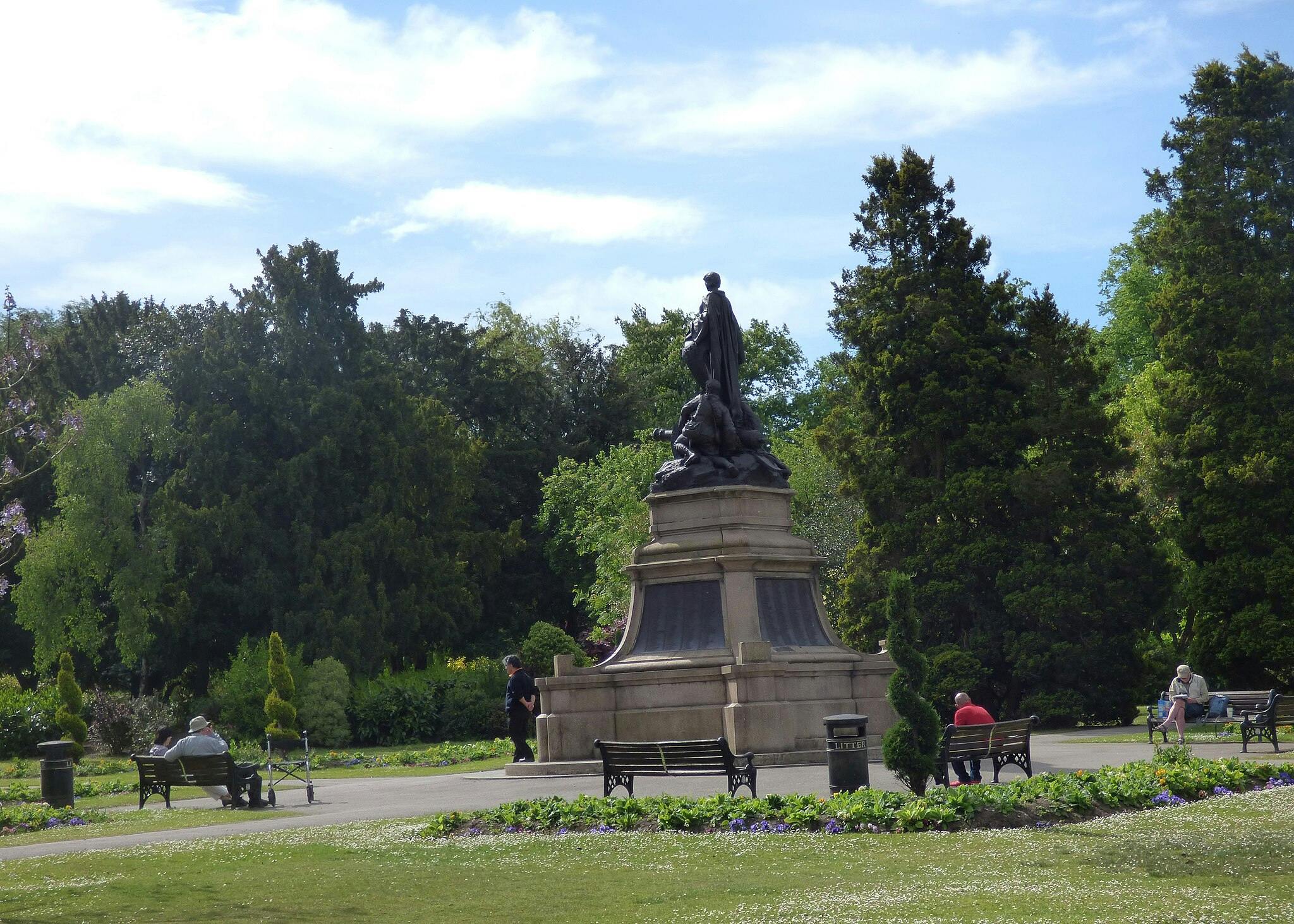 Une statue dans un parc entouré d'arbres et de bancs, avec plusieurs personnes assises et se promenant.