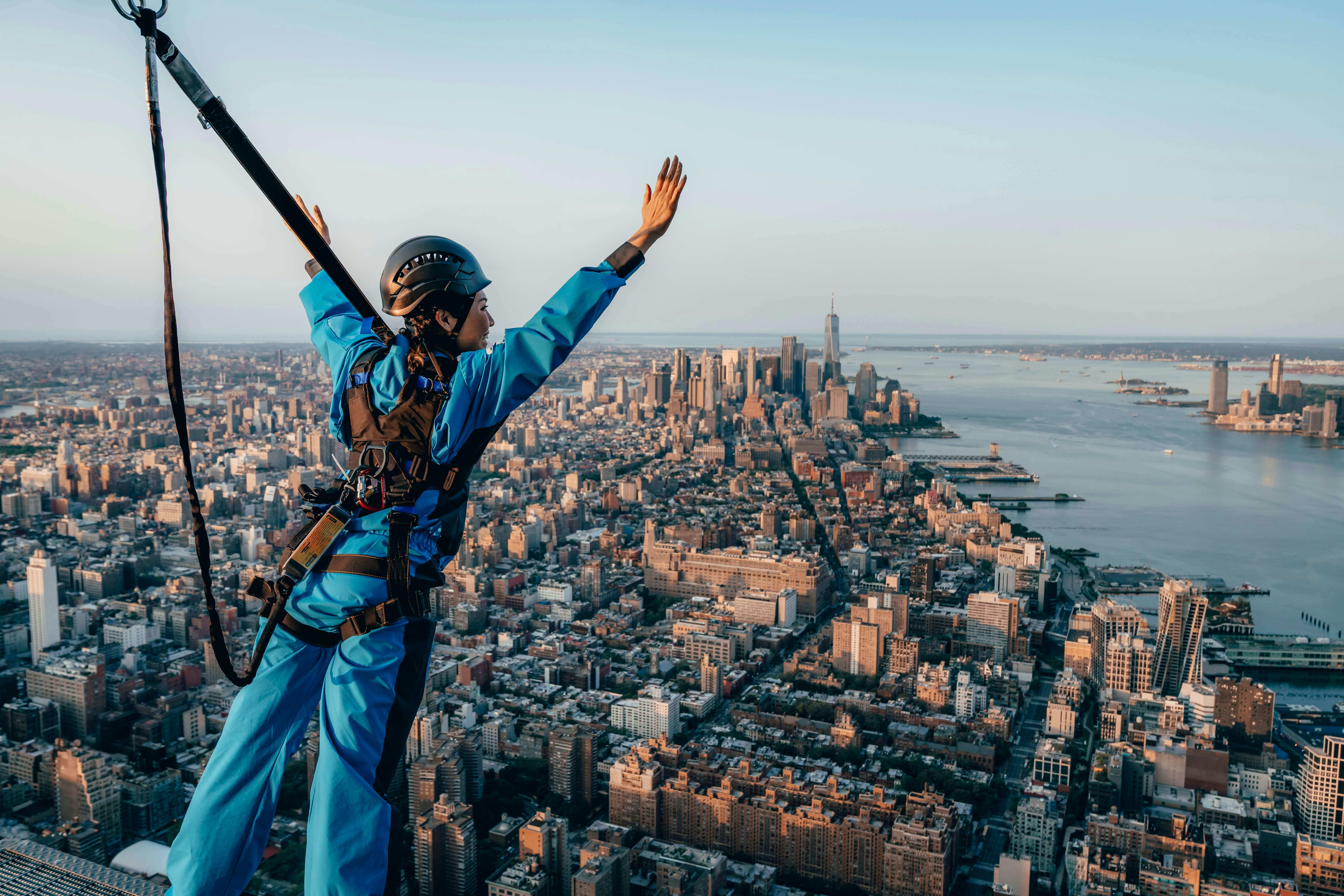 Une personne vêtue d'une combinaison bleue se tient à une altitude élevée, les mains levées, surplombant un paysage urbain tentaculaire et un plan d'eau.