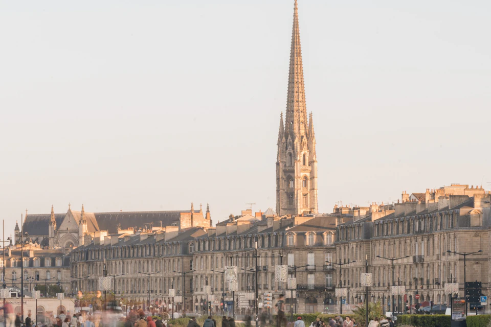 A cityscape featuring historic buildings and a tall church spire under a clear sky, with people in the foreground.