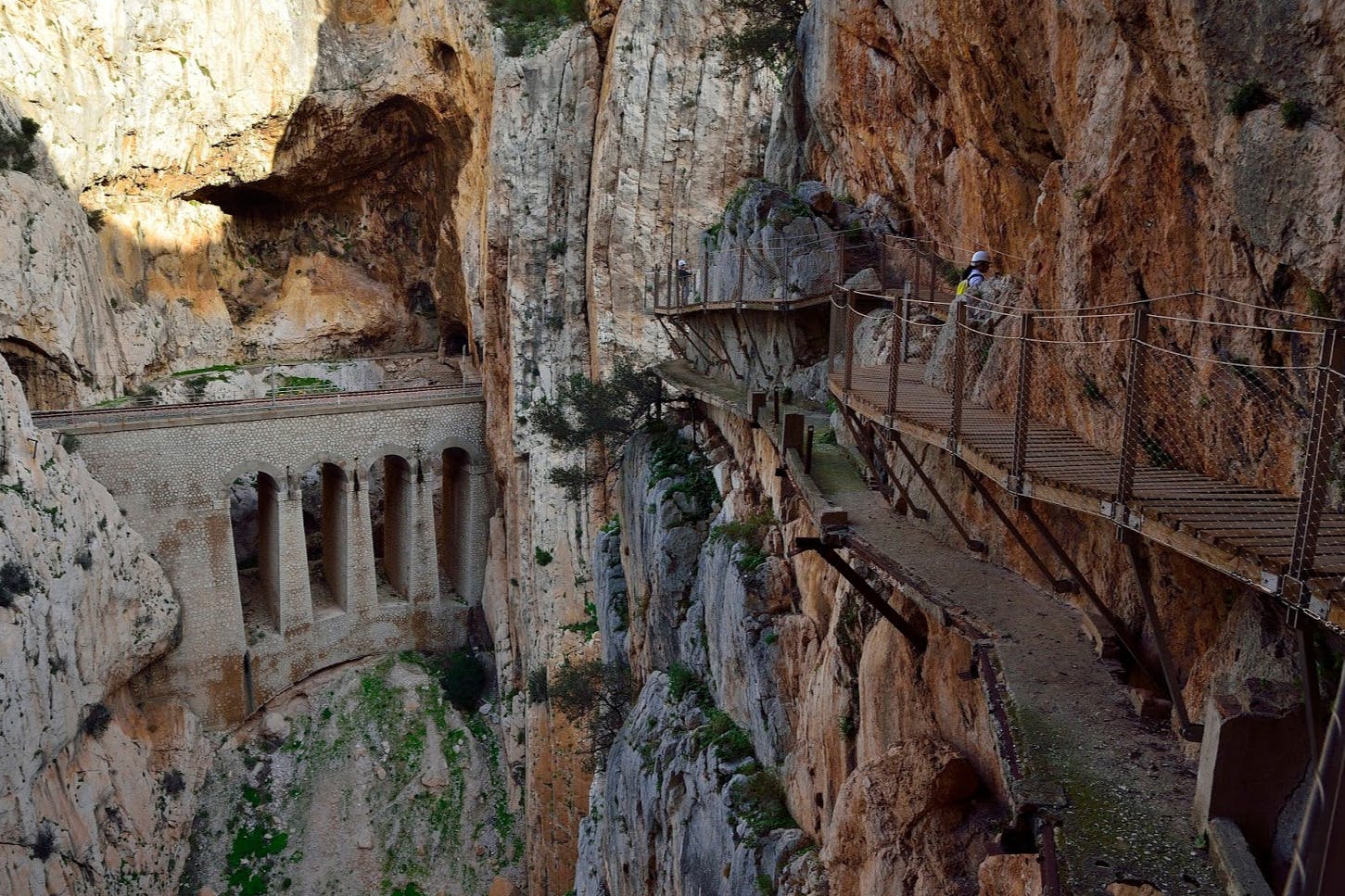 A narrow, elevated walkway clings to a sheer cliff, with a person visible on the path beside a stone bridge spanning a deep gorge.