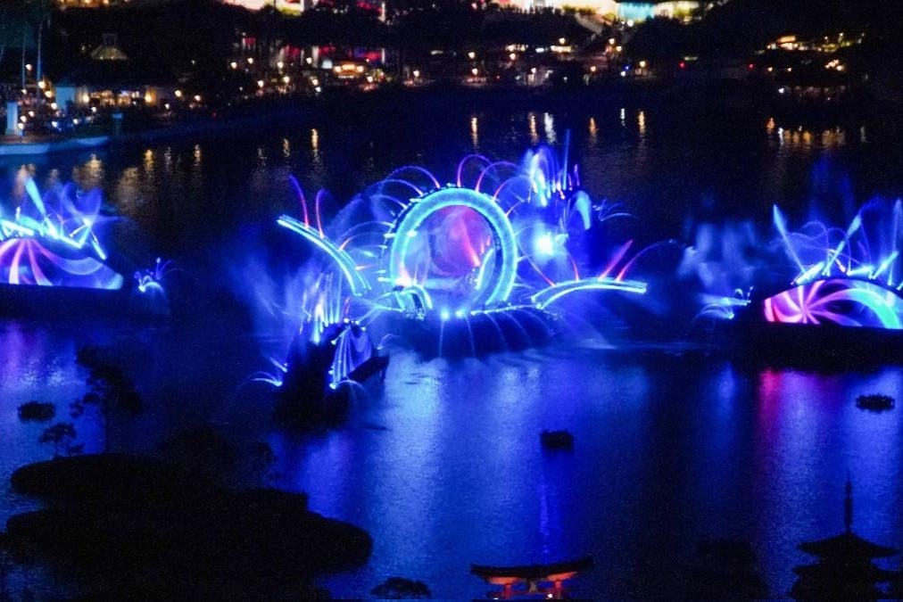 A nighttime water show with illuminated fountains and a large, glowing circular structure surrounded by spray and lights.