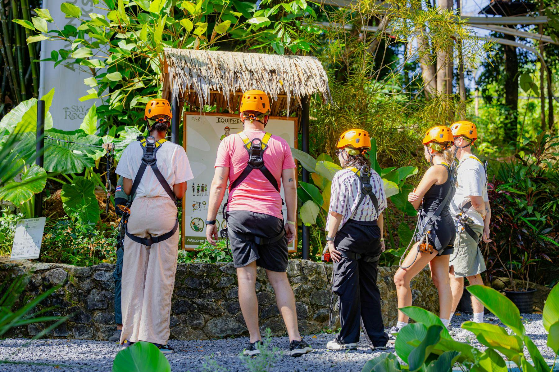A group of people wearing harnesses and orange helmets stand in front of an informational sign in a tropical outdoor setting.