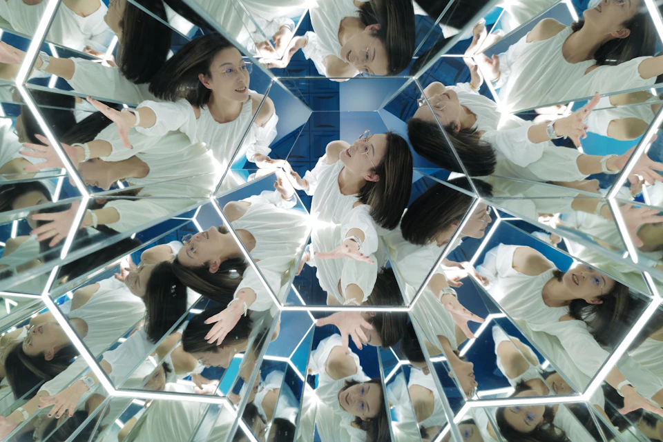 A person in white surrounded by reflective mirrors creating multiple reflections in a geometric pattern.