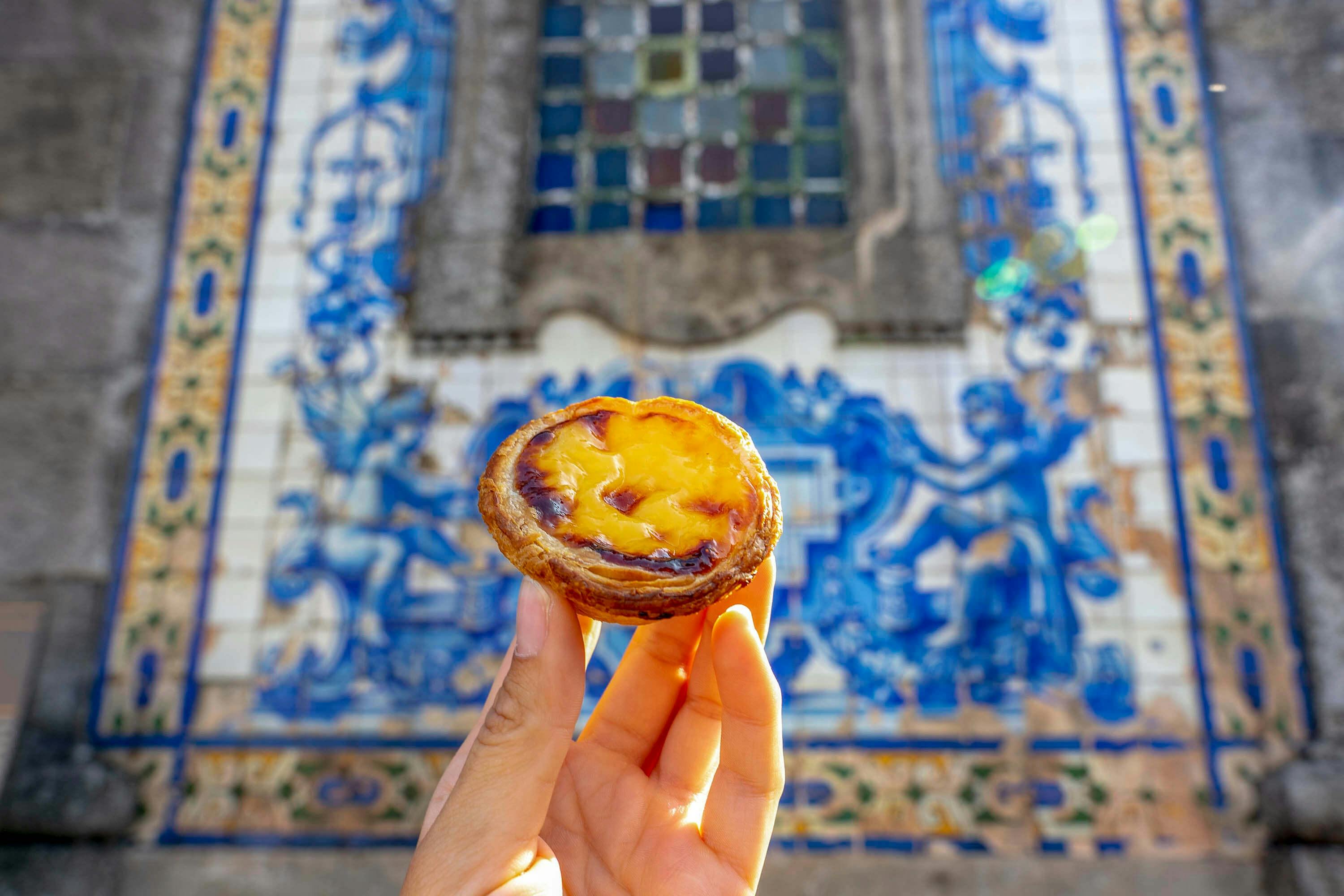 Ruka držící portugalský pudinkový koláč před modro-bílou kachlovou nástěnnou malbou s vitrážovým oknem nahoře.