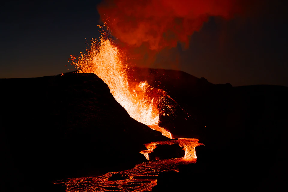 Molten lava erupts and flows from a volcano at night, with bright orange and red hues contrasting a dark sky.